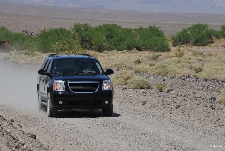 A black GMC Yukon kicks up dust as it drives slowly on a dirt road in a desert.