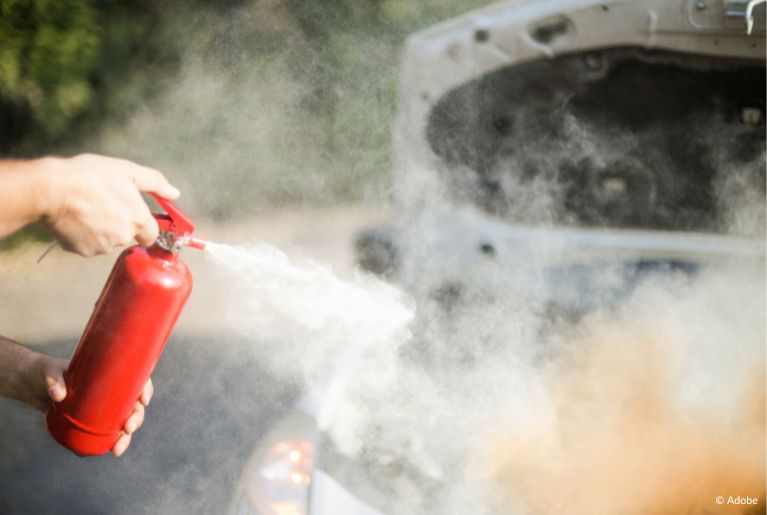 A person uses a fire extinguisher to subdue the flames from the engine compartment of a white car.