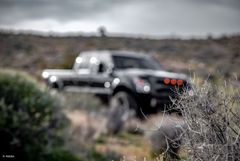 A black Ford F-150 truck is out of focus, parked in a shrubby desert field.