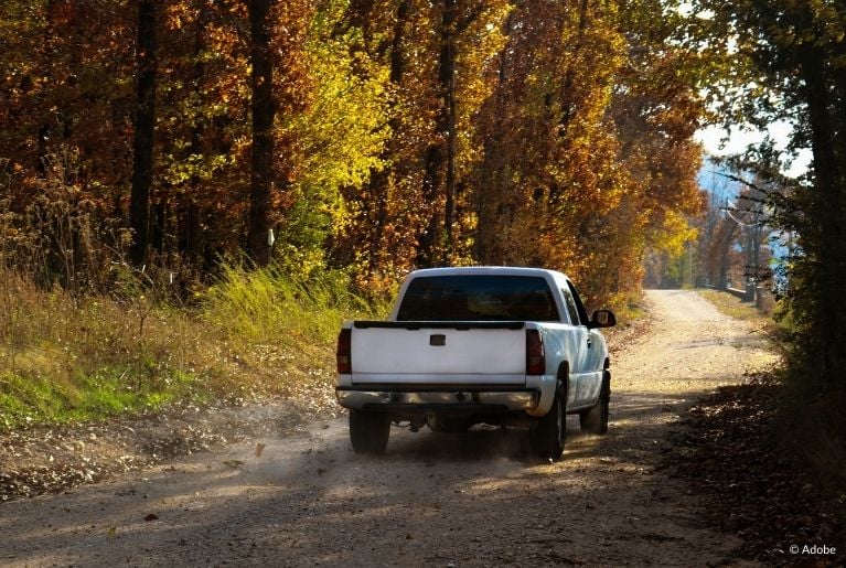 A white pickup truck drives on a dirt round surrounded by trees. The color of the leaves indicates that it is fall.