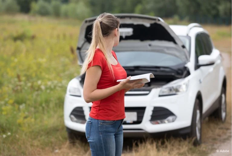 A woman in a red shirt stands in front of her white SUV. She is holding an owner's manual. The white SUV has its hood popped open.