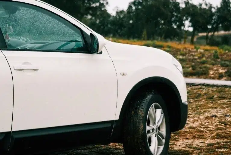 A white Nissan Murano vehicle is parked in a muddy, grassy field beside an empty street.