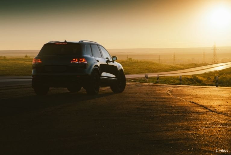 A GMC Terrain SUV is parked on an empty stretch of concrete. It is sunset. The orange sunlight illuminates a long stretch of highway on otherwise undeveloped land.