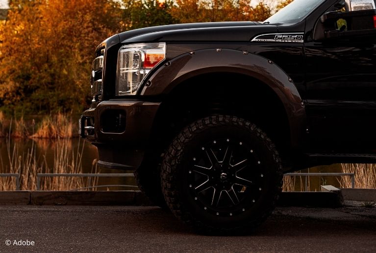 A close-up of the front of a Ford F-250 pickup truck. The black truck is parked on the side of the road, next to a river in fall.