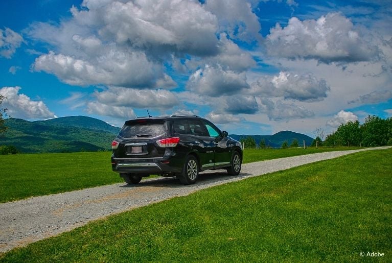 A black Nissan Pathfinder is parked on a single lane. The lane bisects a grassy field. A blue sky is littered with clouds.