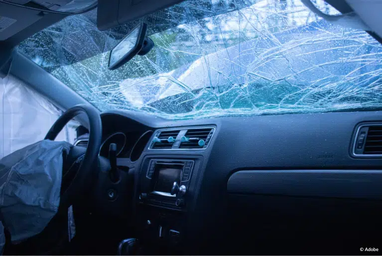 An interior view of a vehicle post-crash. The frontal hood is crumpled open, the windshield is cracked, and the frontal and side airbags have deployed.