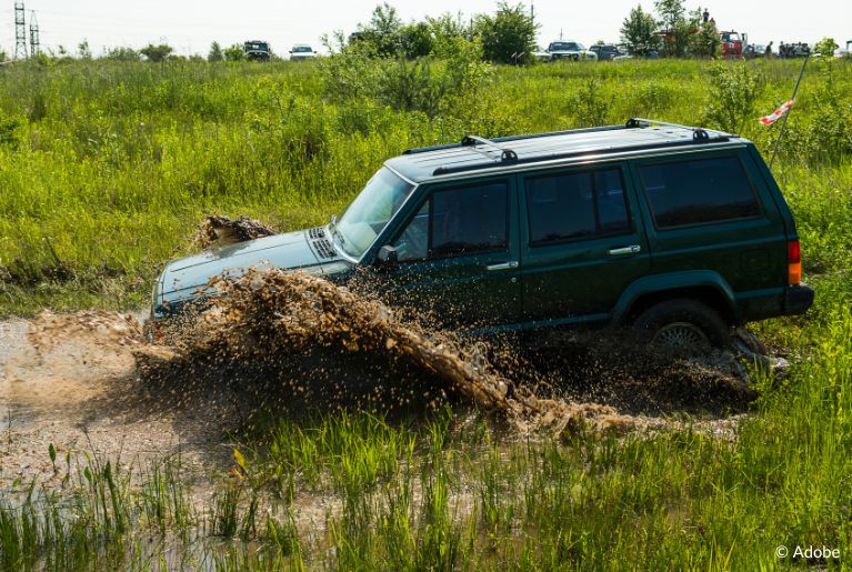 A dark green FCA SUV drives into a muddle pool in the middle of a grassy wetland.