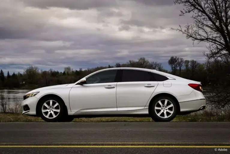 A white sedan is parked on the side of the road, just in front of a river. It is overcast.