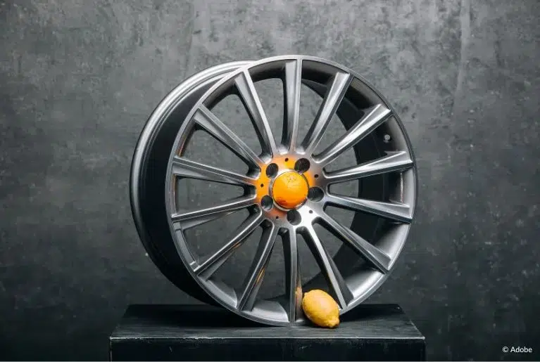 A wheel rim with an orange in the center and a lemon at the bottom. The wheel rim is placed on a black podium.