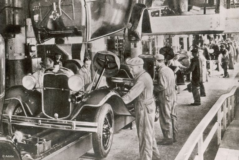 A black and white image of factory workers assembling Ford Model T vehicles.