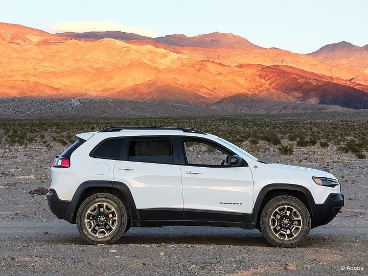 A white Jeep Cherokee SUV is parked in the middle of a desert, with shrubs and hillsides in the background.