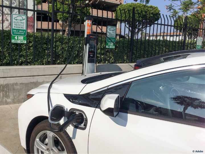 A white electric vehicle is plugged into a charger at an outdoor parking station in a parking lot. The electric vehicle parking spot has a time limit of four hours.