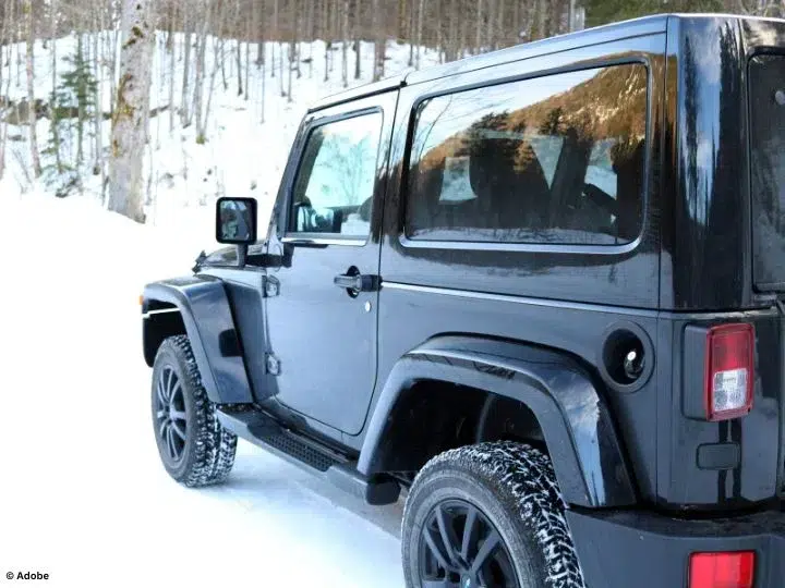 A black Jeep is parked in the snow. In the background are bare, thin trees.