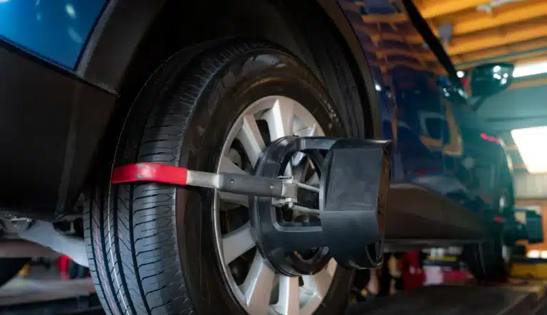 Close-up of a car's wheel undergoing alignment in a garage. A clamp and sensor device is attached to the tire, with the blue car elevated on a lift.