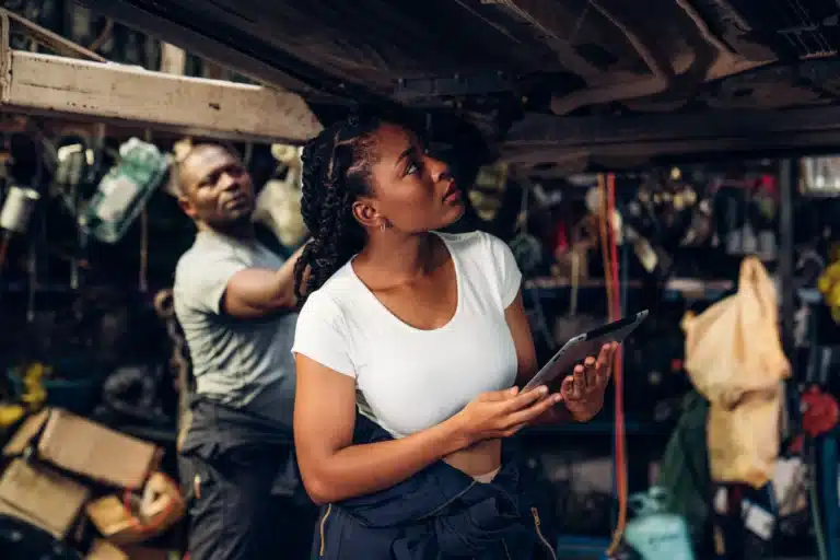 A woman holding a tablet inspects the underside of a vehicle in a garage, while a man works in the background.